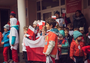 Dzieci stoją na schodach przed przedszkolem, trzymają flagę